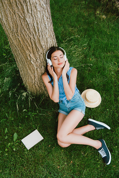 high angle view of beautiful woman in closed eyes listening music while sitting on grass near book and straw hat