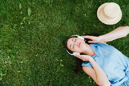 top view of pretty woman with closed eyes lying on grass and listening music near straw hat