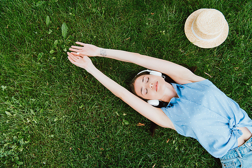 top view of attractive woman lying on grass and listening music near straw hat