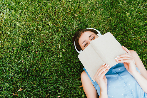 top view of woman in wireless headphones lying on grass and holding book