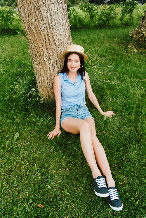 happy girl in straw hat sitting on green grass near tree trunk