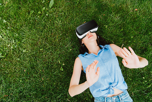 top view of young woman in virtual reality headset gesturing and lying on green grass