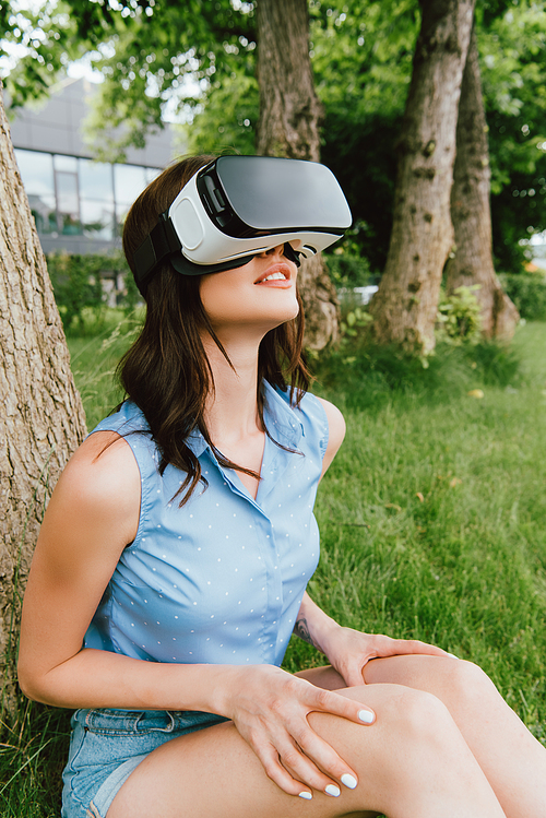 happy young woman in virtual reality headset sitting near green trees