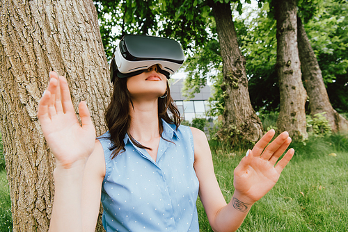 selective focus of young woman in virtual reality headset gesturing near green trees