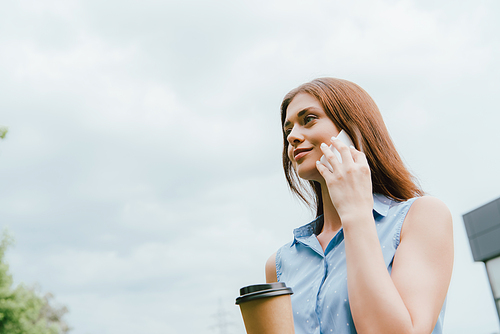 low angle view of businesswoman holding paper cup and talking on smartphone against cloudy sky