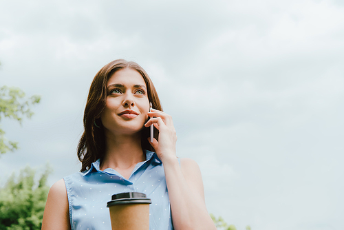 low angle view of businesswoman holding disposable cup and talking on smartphone against cloudy sky