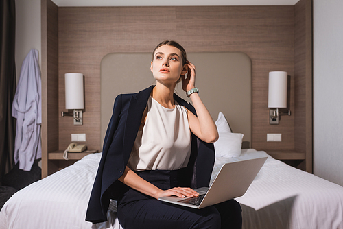 woman in suit sitting on bed with laptop and touching hair in hotel room