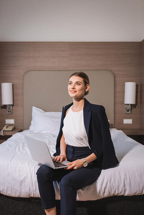 dreamy woman in suit sitting on bed with laptop in hotel room