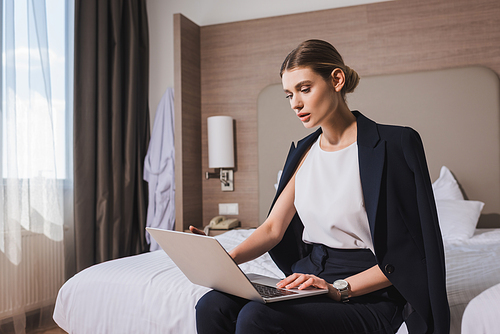 woman in suit sitting on bed and looking at laptop in hotel room
