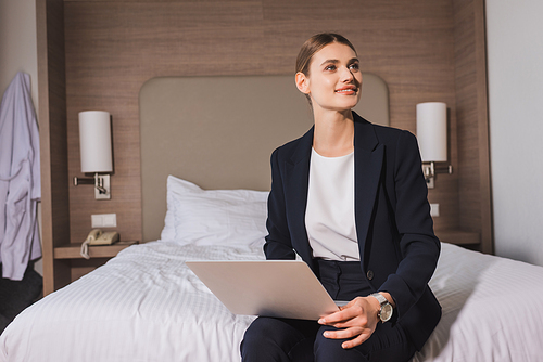 joyful woman in suit sitting on bed with laptop and looking away in hotel room