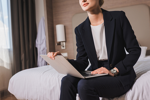cropped view of woman in suit sitting on bed with laptop in hotel room