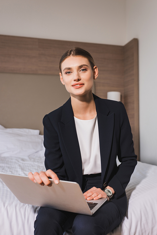 joyful woman in suit sitting on bed with laptop and  in hotel room