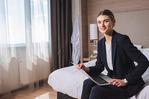 pleased young woman in suit sitting on bed with laptop in hotel room