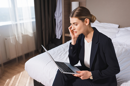 businesswoman in suit sitting on bed and looking at laptop in hotel room