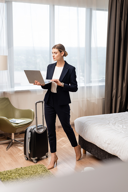 young businesswoman standing and using laptop near travel bag in hotel room