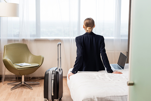 back view of businesswoman sitting on bed near laptop with blank screen and suitcase in hotel room