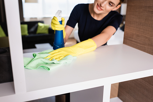 selective focus of maid in rubber gloves holding spray bottle and rag while cleaning shelf in hotel room