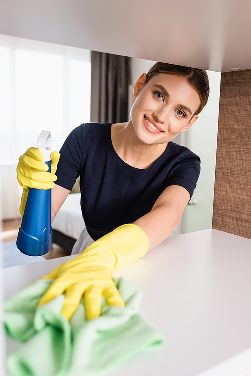 selective focus of joyful maid in rubber gloves holding spray bottle and rag while cleaning shelf in hotel room
