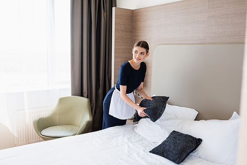 joyful chambermaid in apron holding pillow near bed in hotel