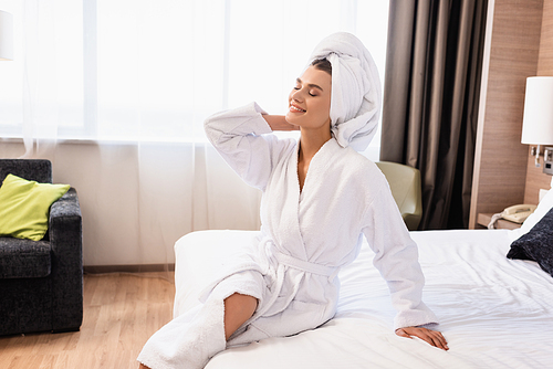 joyful woman with closed eyes in white towel and bathrobe sitting on bed in hotel room