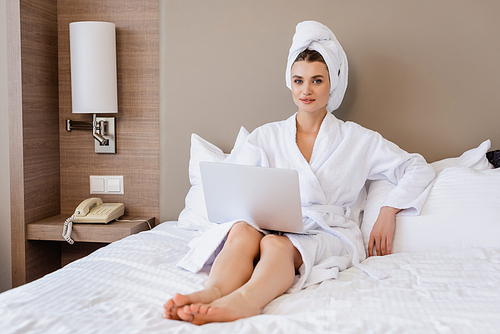 selective focus of barefoot young woman in bathrobe using laptop on bed