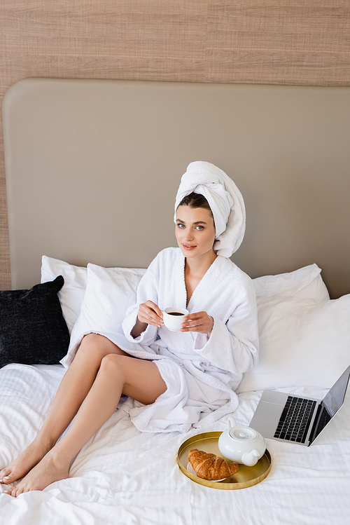 young woman in bathrobe holding cup of coffee near laptop and breakfast tray on bed