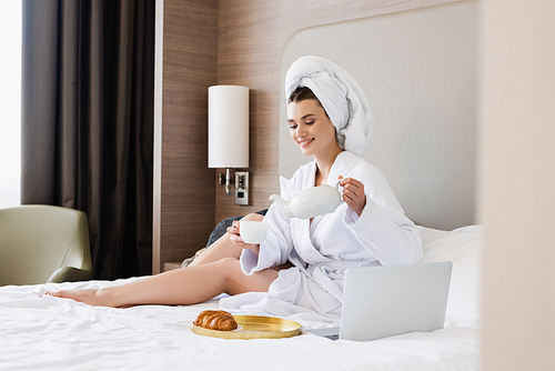 woman in bathrobe holding cup and teapot near laptop and tray with breakfast on bed