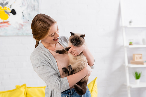 Cheerful woman holding siamese cat at home