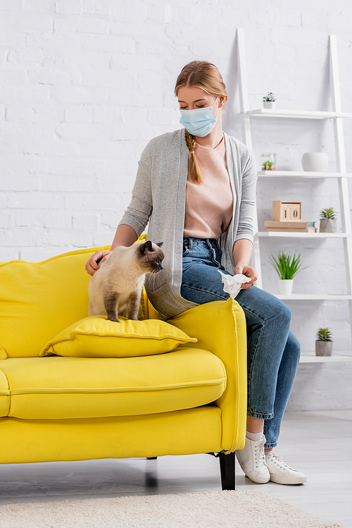 Young woman in medical mask holding napkin near cat on couch