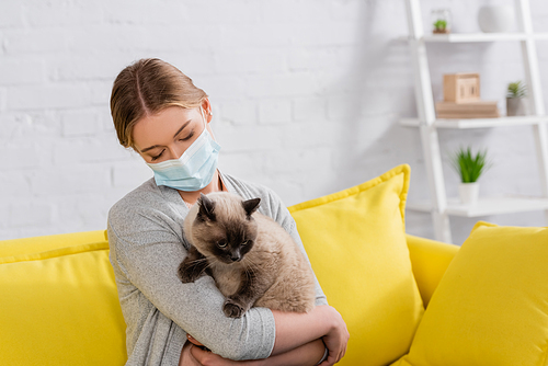 Woman in medical mask holding siamese cat during allergy in living room