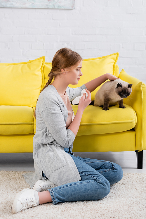 Young woman with napkin sitting on carpet near siamese cat on couch
