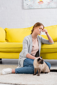 Young woman with napkin feeling allergy reaction near siamese cat on carpet