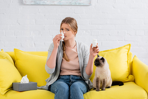 Woman with allergy holding napkin and pills near siamese cat on couch