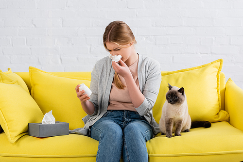 Woman with allergy holding napkin during snuffle and pills near furry siamese cat