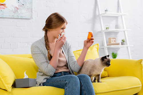 Young woman holding pills and napkin during allergy reaction near siamese cat on bright couch