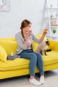 Woman with allergy looking at jar with pills near cat on yellow couch at home