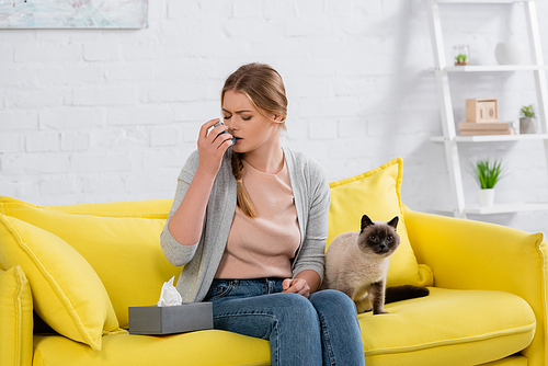 Woman with allergy using inhaler near furry siamese cat on sofa