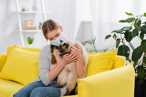 Young woman with allergy reaction wearing medical mask and holding furry cat on yellow couch