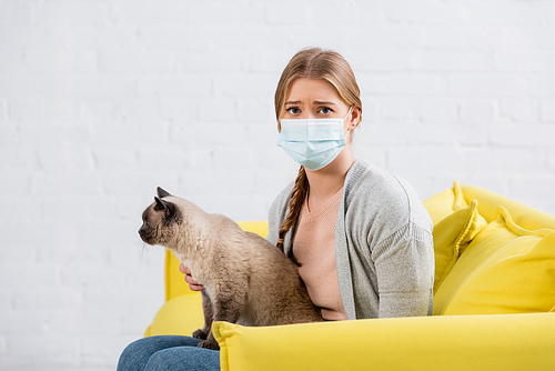 Woman with allergy wearing medical mask and petting siamese cat on yellow couch