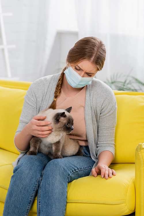 Sad woman in medical mask looking at siamese cat during allergy on bright couch