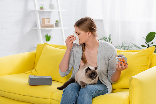 Woman with allergy holding pills and napkin near furry siamese cat on yellow sofa