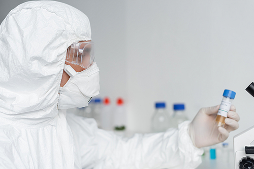 Scientist in protective suit and goggles holding vaccine on blurred background