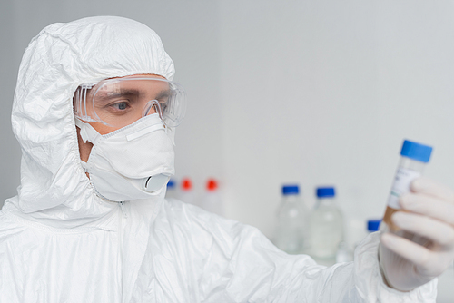 Scientist in protective suit and mask holding vaccine on blurred foreground