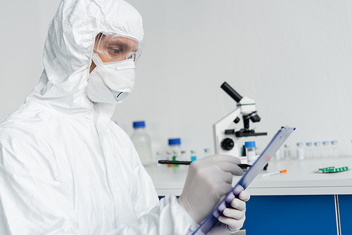 Scientist in protective uniform writing on clipboard on blurred foreground