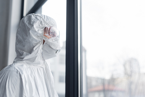 Scientist in medical mask, goggles and hazmat suit standing near window