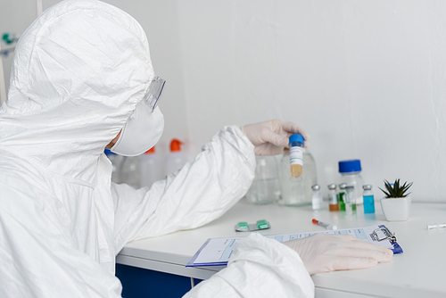 Scientist in protective uniform and latex gloves holding vaccine near clipboard on blurred foreground