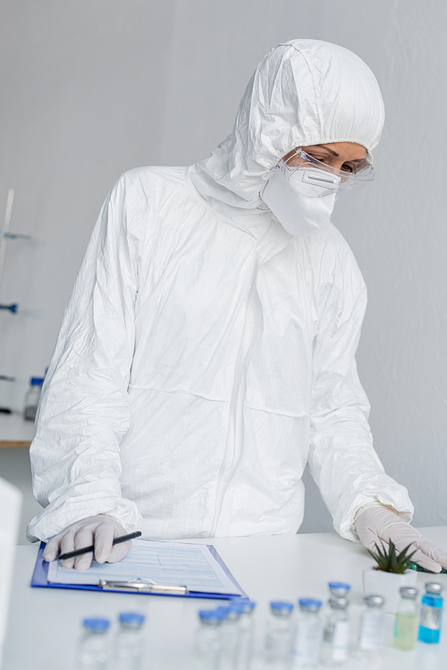 Scientist in hazmat suit standing near vaccines and clipboard on blurred foreground in laboratory
