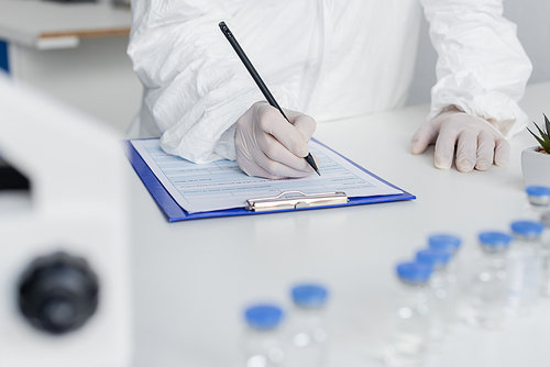 Cropped view of scientist writing on clipboard near vaccines on blurred foreground