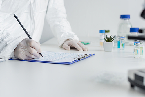 Cropped view of scientist in hazmat suit and latex gloves writing on clipboard near vaccines on blurred foreground