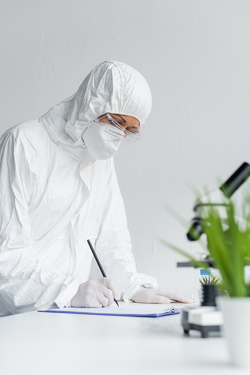 Scientist in protective suit writing on clipboard near microscope on blurred foreground